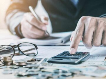 A person calculating and writing on book and glass, coins on table